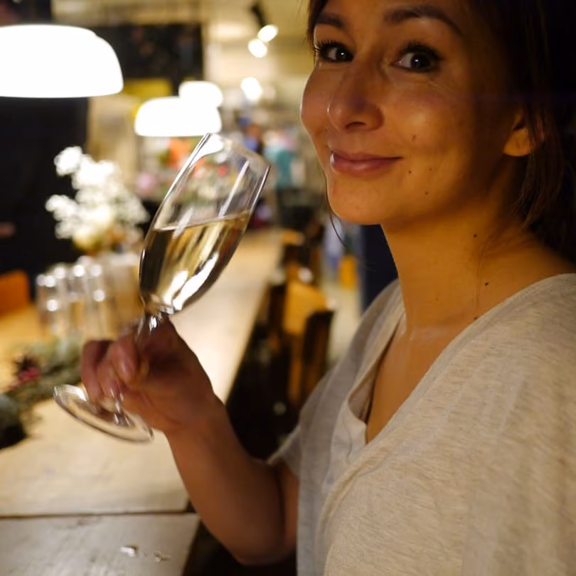 Woman smiling and holding a glass of white wine at a warmly lit wooden bar.