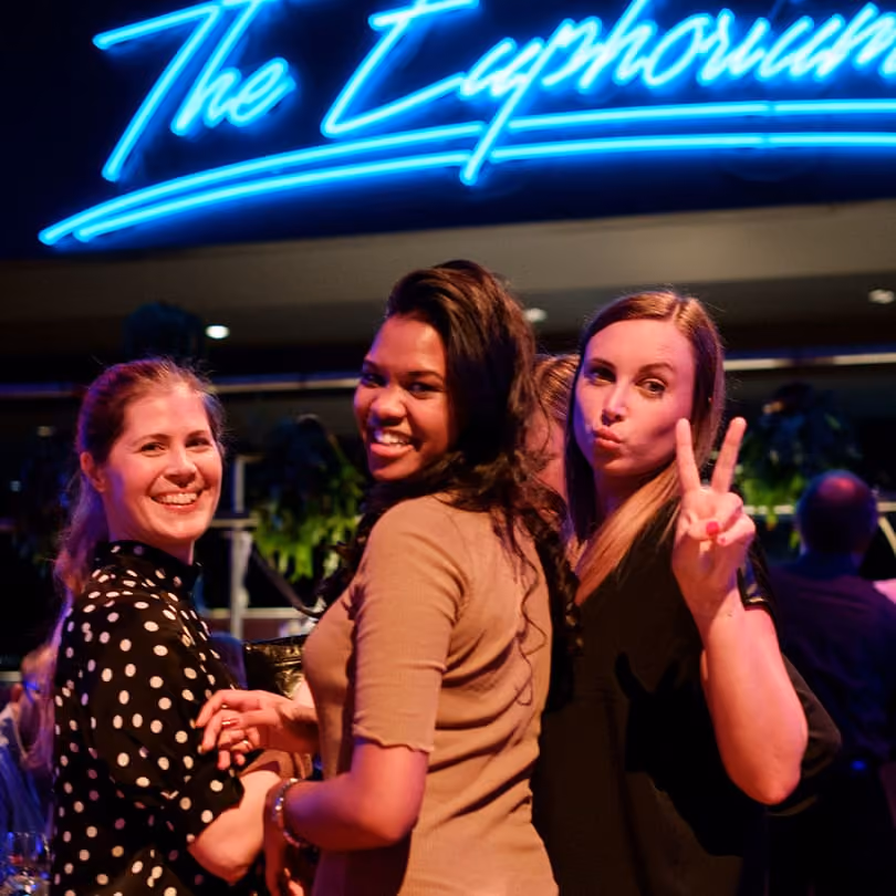 Three women smiling and posing at night under a neon sign that reads The Euphorium.