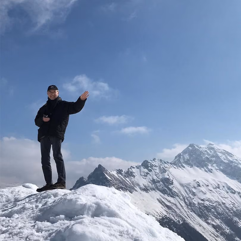 Person in black jacket and cap waving while standing on snow-covered mountain peak with blue sky.