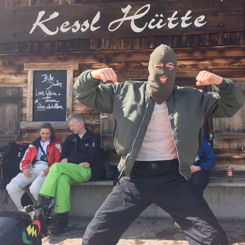 Person wearing green balaclava and jacket posing with flexed arms in front of wooden Kessl Hütte hut.