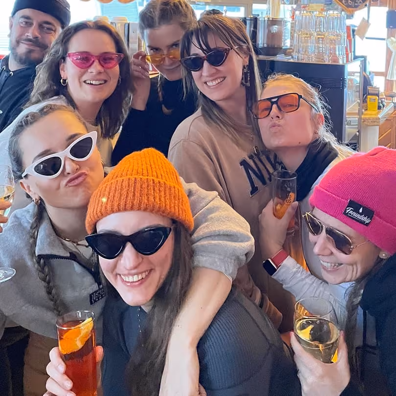 Group of seven smiling friends wearing colorful sunglasses and holding drinks in a lively indoor setting.