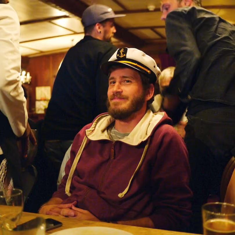 Bearded man in a captain's hat and maroon hoodie sitting at a wooden table smiling.