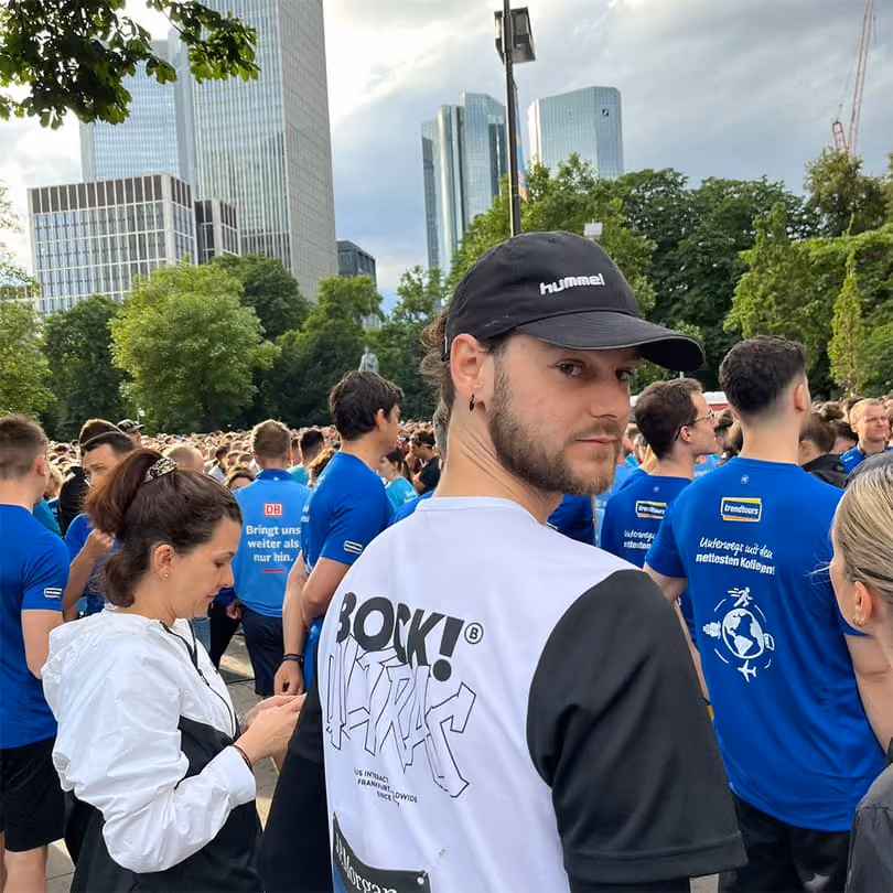 Man in white and black shirt looks at camera in a crowd of people wearing blue shirts outdoors.