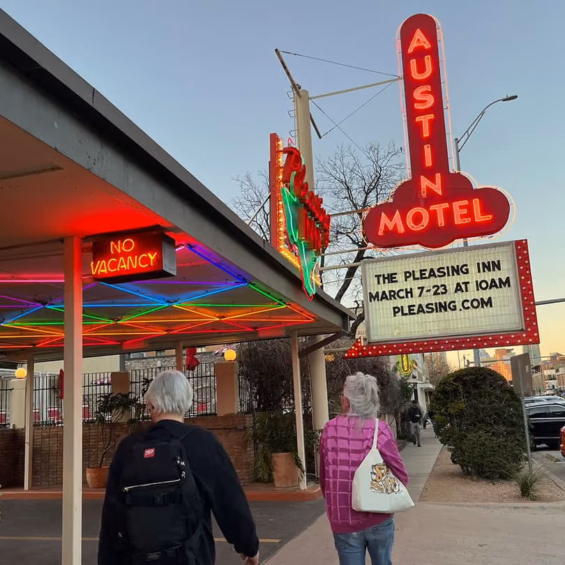 Two people walk near the neon-lit Austin Motel sign with a no vacancy sign and an event announcement.