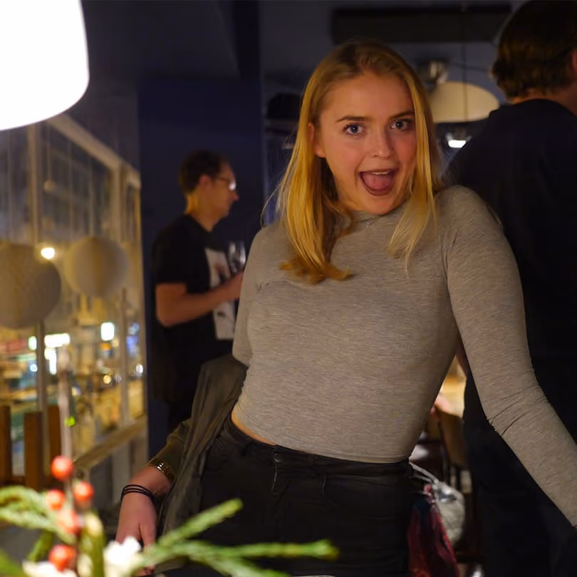 Young woman with blonde hair and grey top posing excitedly indoors with people and lights behind her.