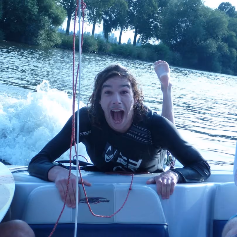 Man with long hair and wetsuit excitedly holding the back of a boat on a river with trees in background.