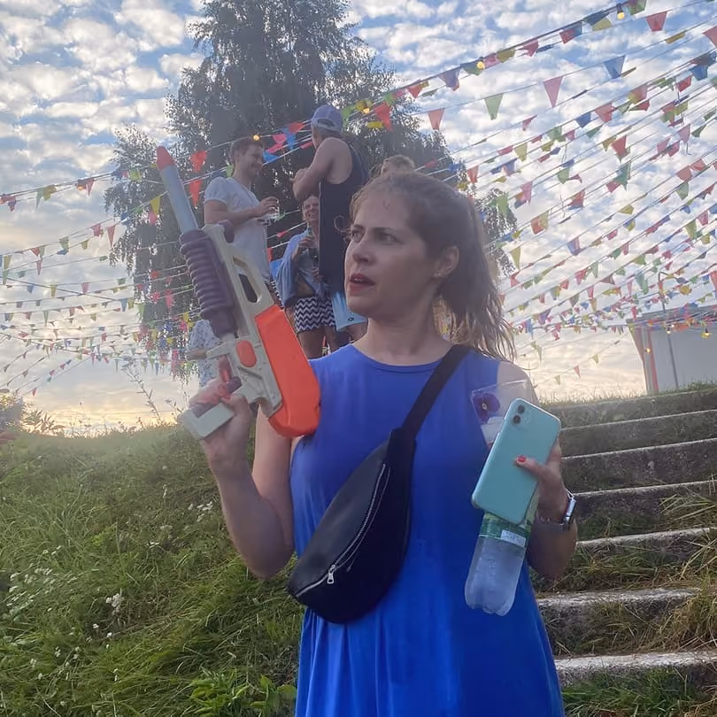 Woman in blue dress holding a toy water gun, phone, and bottle with festive flags and people behind.