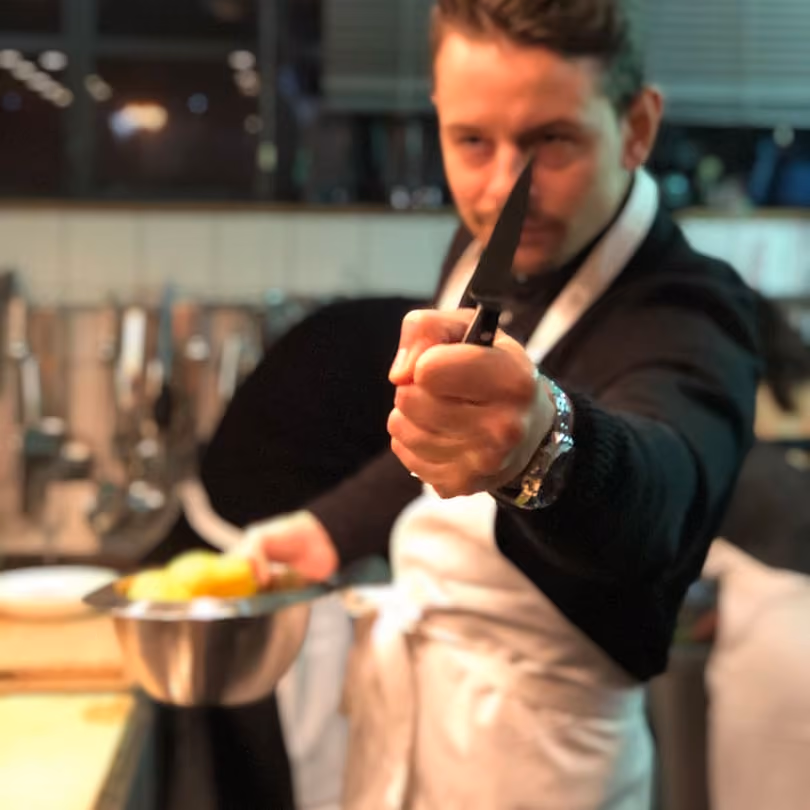 Chef in a kitchen holding a knife forward with one hand and a bowl with food in the other hand.