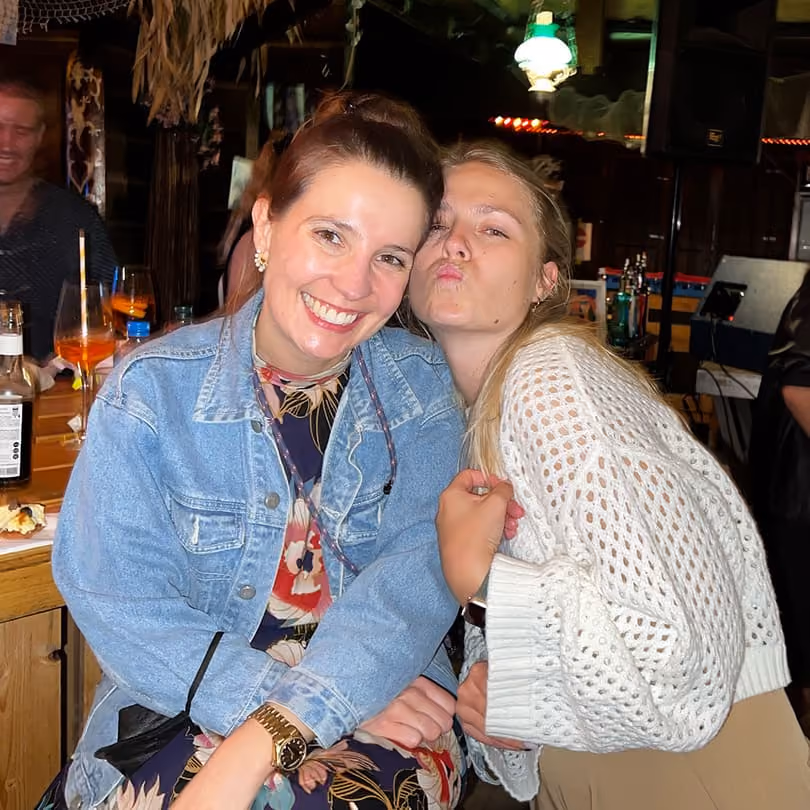 Two women smiling and posing closely in a dimly lit bar with drinks and wooden decor.