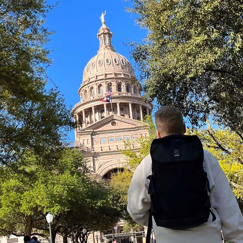 Person wearing a black backpack walking toward the Texas State Capitol in Austin with trees on sides.