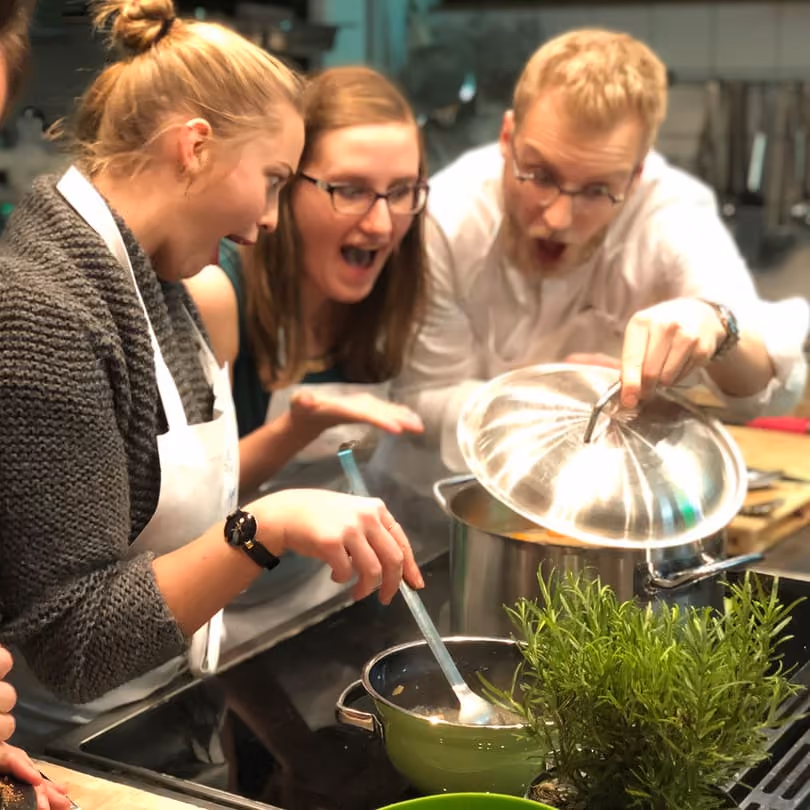 Three people excitedly look inside a pot as one lifts the lid in a kitchen setting.