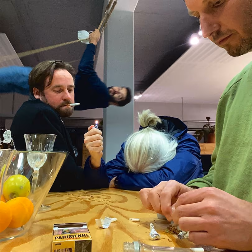 Four people around a table, one lighting a cigarette, one rolling tobacco, one with head down, and one swinging behind.