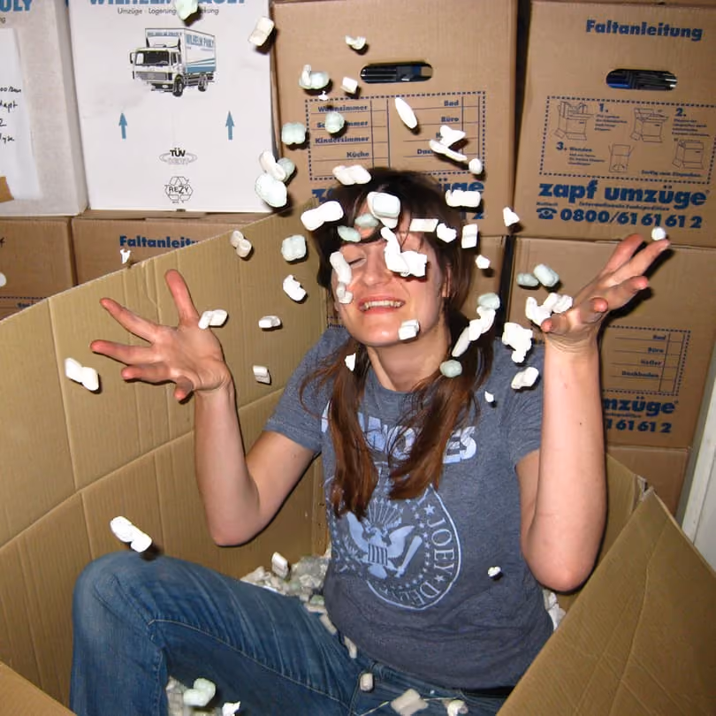 Person sitting in a cardboard box throwing packing peanuts in the air with eyes closed and smiling.