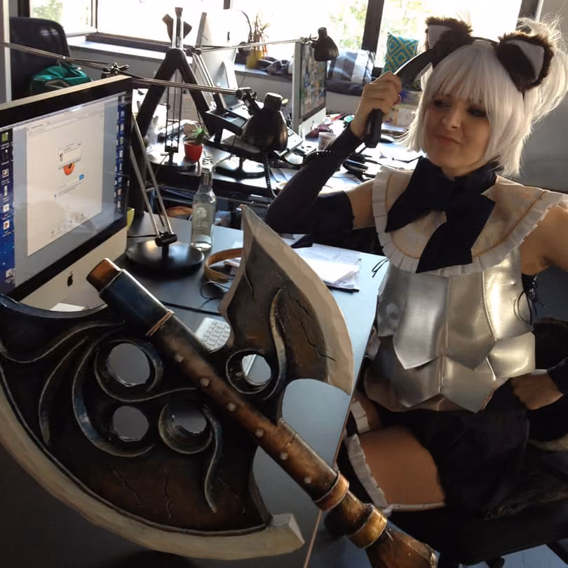 Person in cat-eared costume holding a large fantasy battle axe in an office with computers and desks.