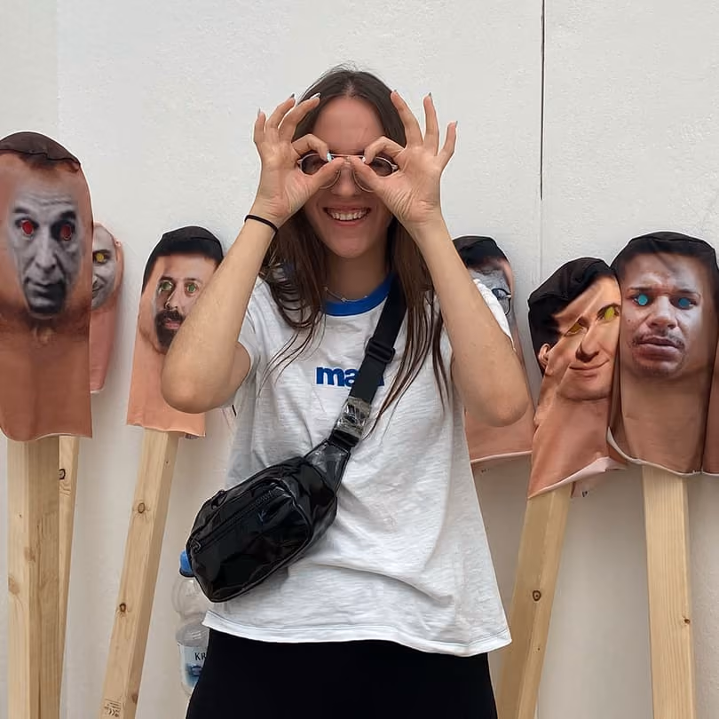 Smiling woman making glasses shape with her hands in front of faces printed on peach-colored masks on sticks.