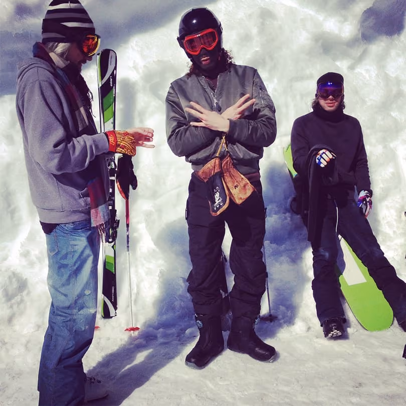 Three people dressed in winter gear standing on snow with skis and a snowboard behind them.