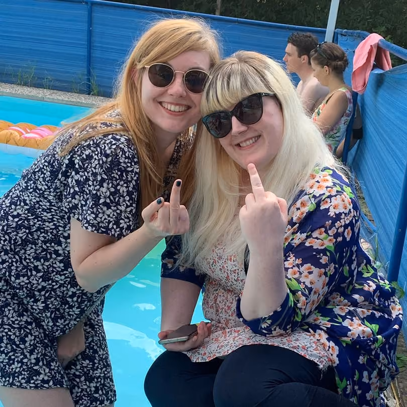 Two smiling women wearing sunglasses by a pool, both raising their middle fingers playfully.