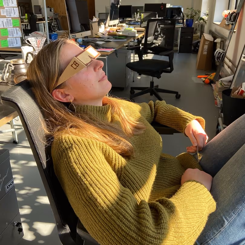 Woman reclining in chair wearing cardboard solar eclipse glasses indoors with office desks around.