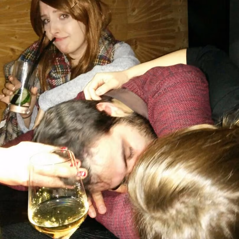 Three people at a party: a woman holds a drink and watches two men kissing closely.
