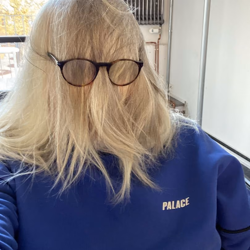 Person with long blonde hair covering their face and wearing glasses and a blue Palace sweatshirt.