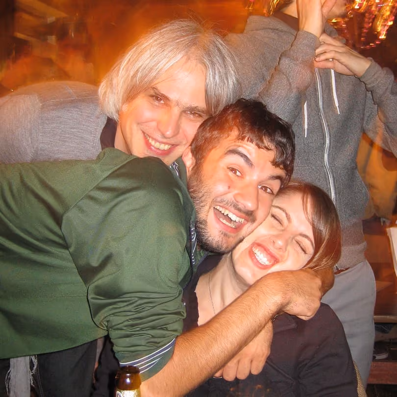 Three friends smiling and hugging closely in a warm, lively indoor setting.
