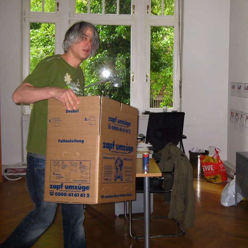 Man with gray hair in green shirt carrying a cardboard box in a room with a window and office chair.