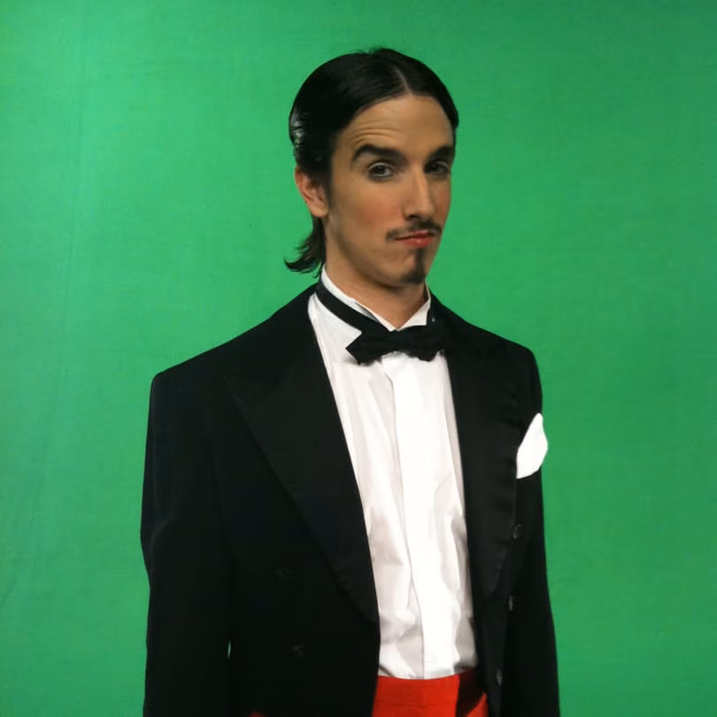 Man with slicked-back hair and mustache in black tuxedo and red pants stands against a green screen background.