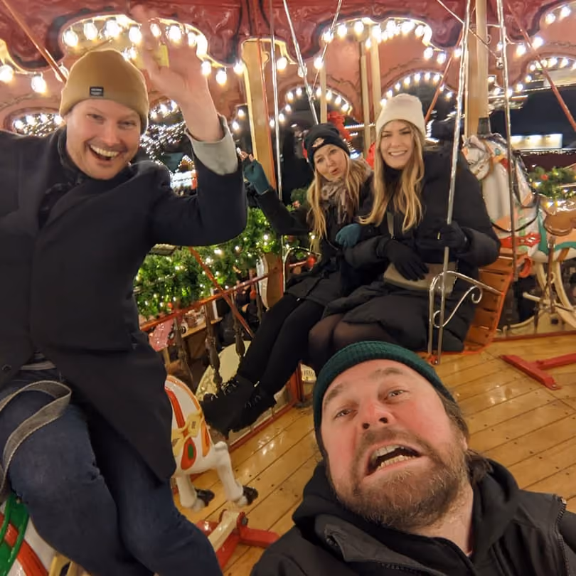 Four people smiling and waving while riding a brightly lit carousel at night.