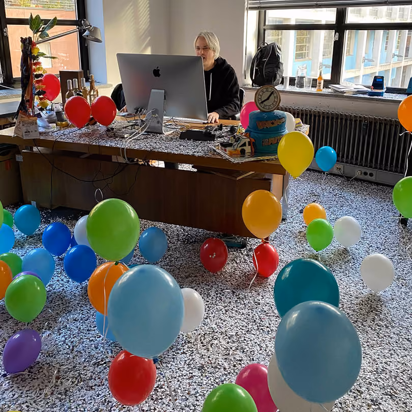 Person working at a desk surrounded by colorful balloons scattered on the carpeted floor.