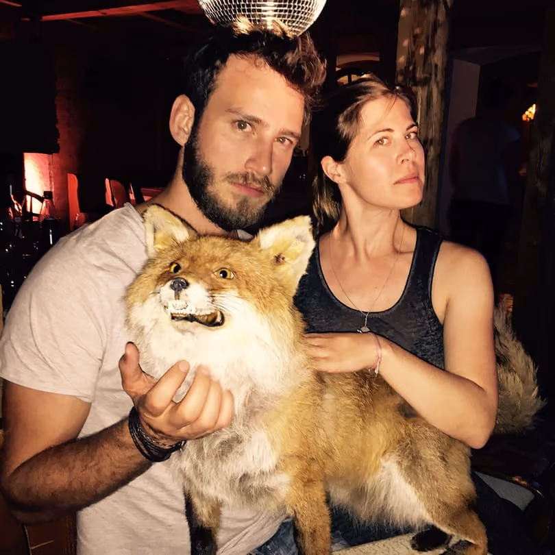 Man and woman posing indoors with a taxidermy fox, with a disco ball overhead.