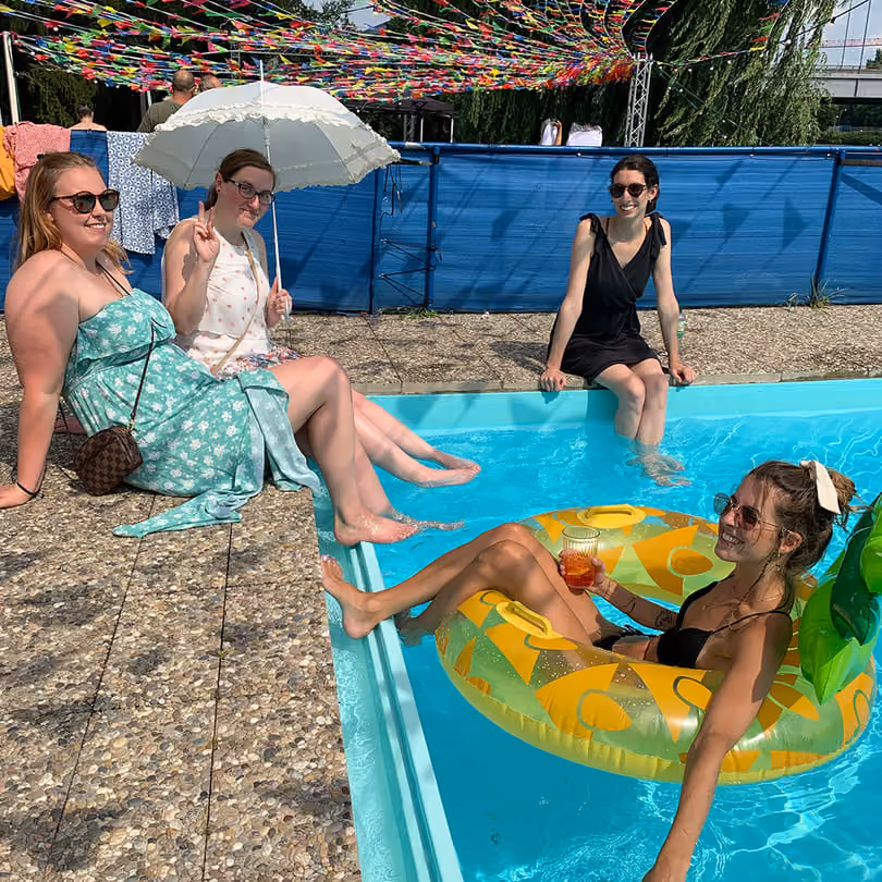 Four women relaxing by a pool; one in a float with a drink, others sitting with feet in water under colorful streamers.