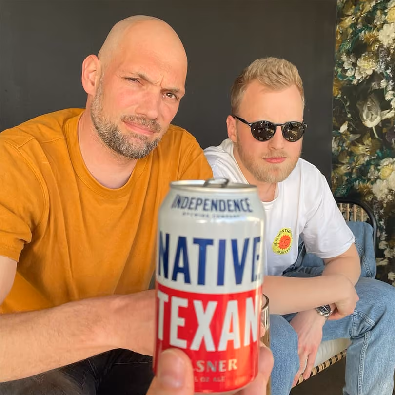 Two men sitting behind a can of Native Texan Pilsner beer held close to the camera.