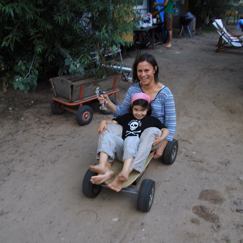 A woman and child sitting and smiling on a homemade wooden go-kart outdoors on dirt ground.