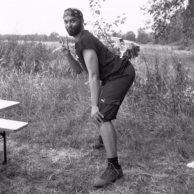 Man outdoors bending forward with hands on knees, flashing peace sign, wearing a bandana and sportswear.