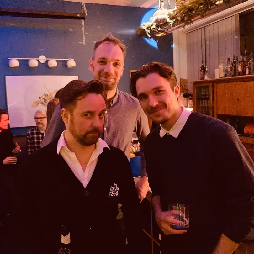 Three men posing in a warmly lit indoor setting, with drinks in hands and others seated in the background.