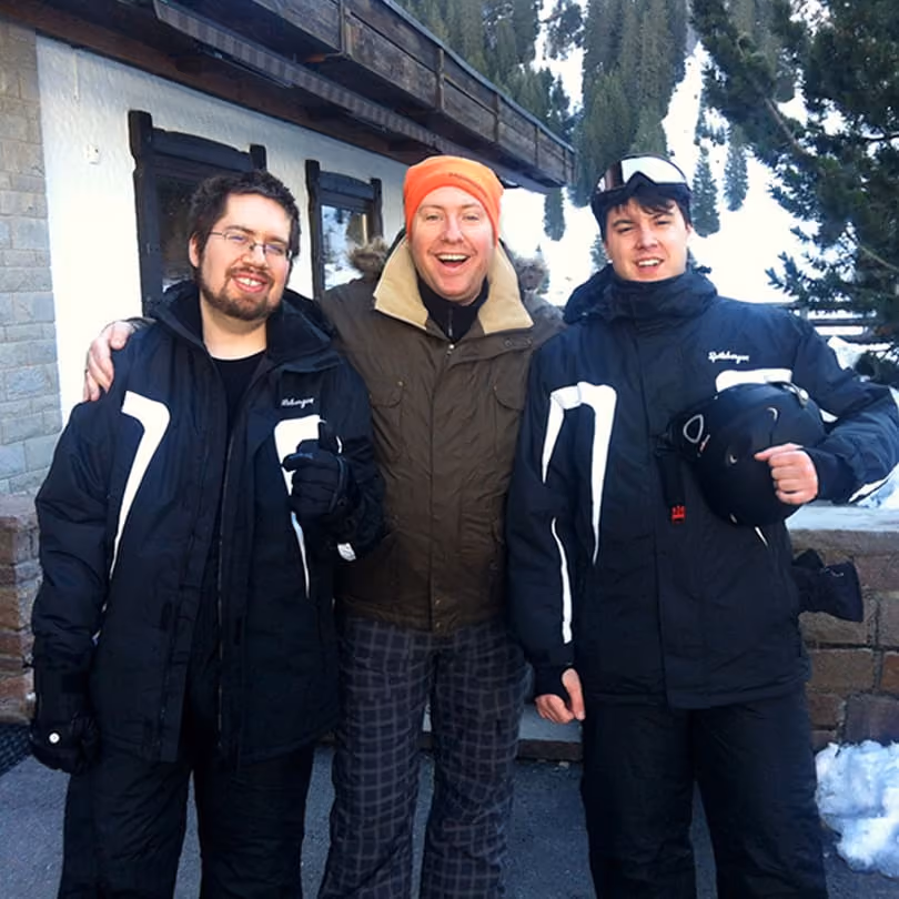 Three men in winter jackets posing with snow and pine trees in the background.
