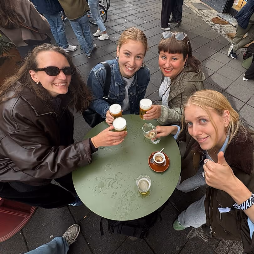 Four smiling women sitting at a round table outdoors, holding glasses of beer and a coffee cup.