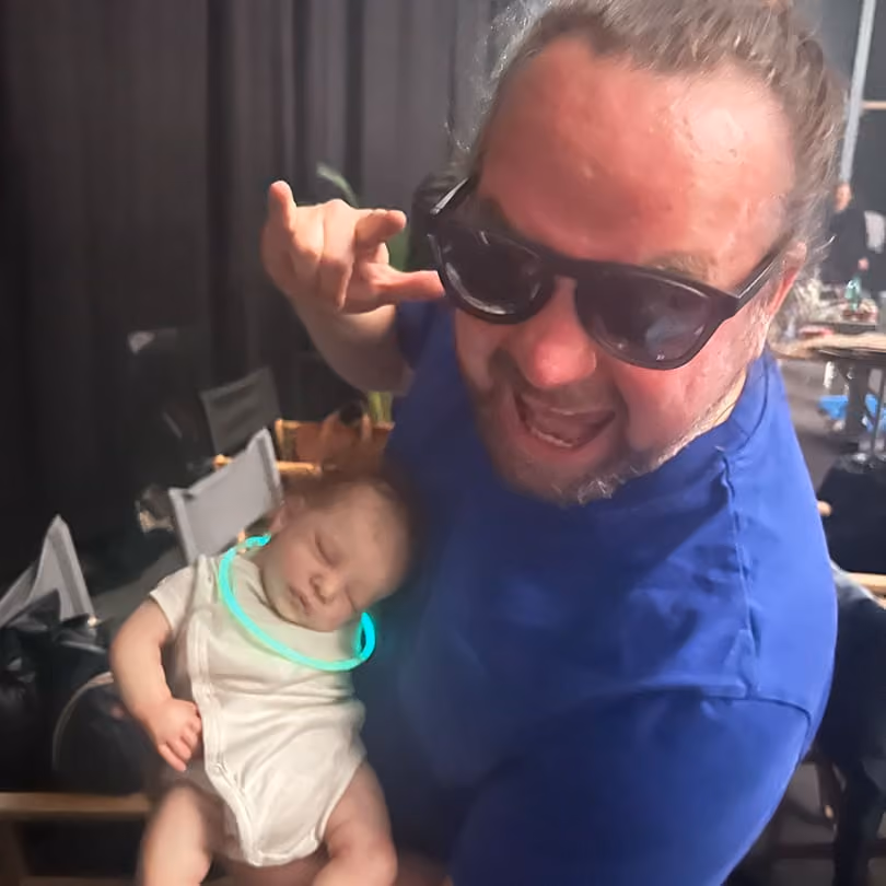 Man wearing sunglasses holding a sleeping baby with a glowing necklace in a dim room.