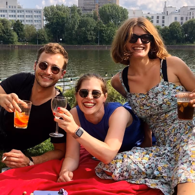Three smiling adults with sunglasses sitting by a river, holding drinks, on a red blanket outdoors.