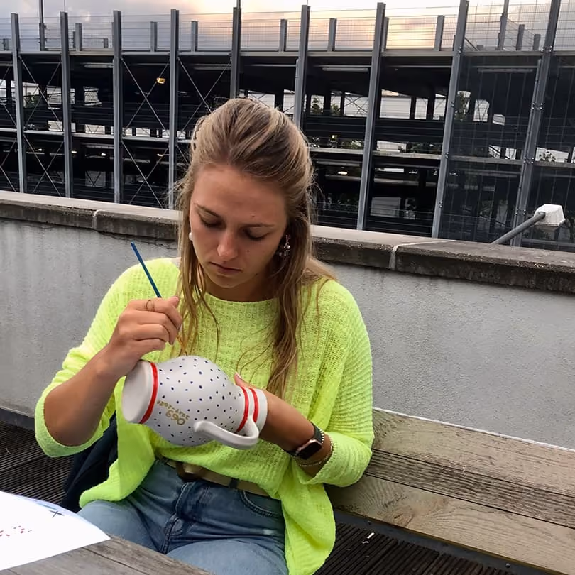 Woman in neon green sweater painting a white ceramic mug with red stripes while seated outdoors.
