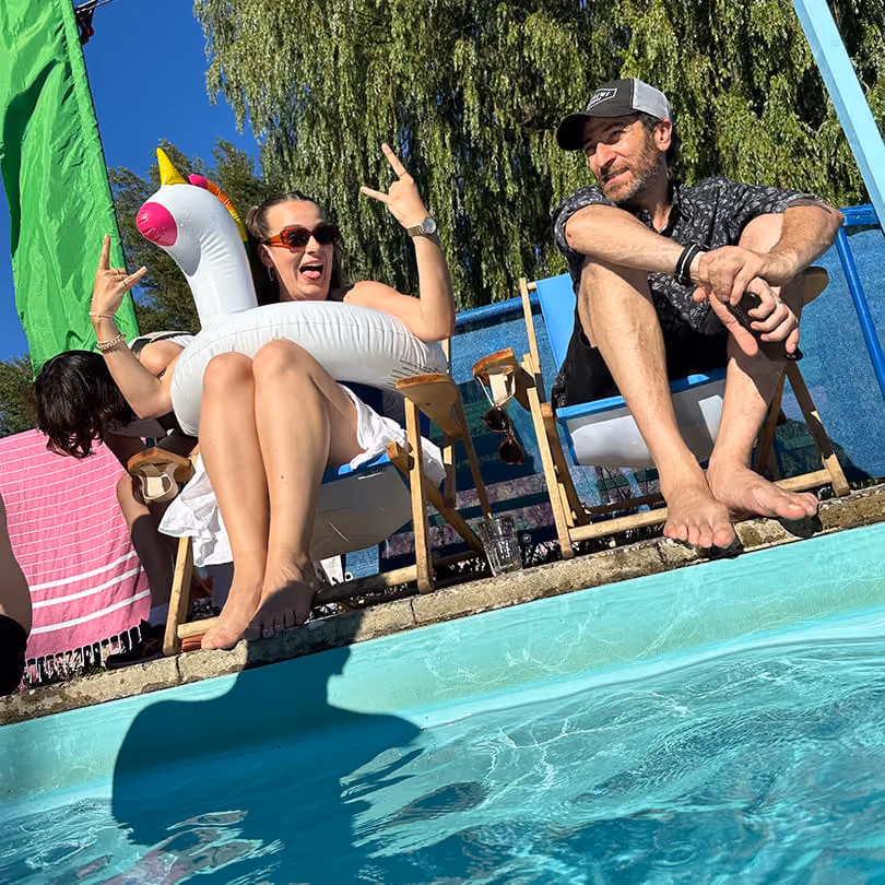 Two people sitting by a pool; woman with swan float making rock signs, man in cap sitting barefoot.