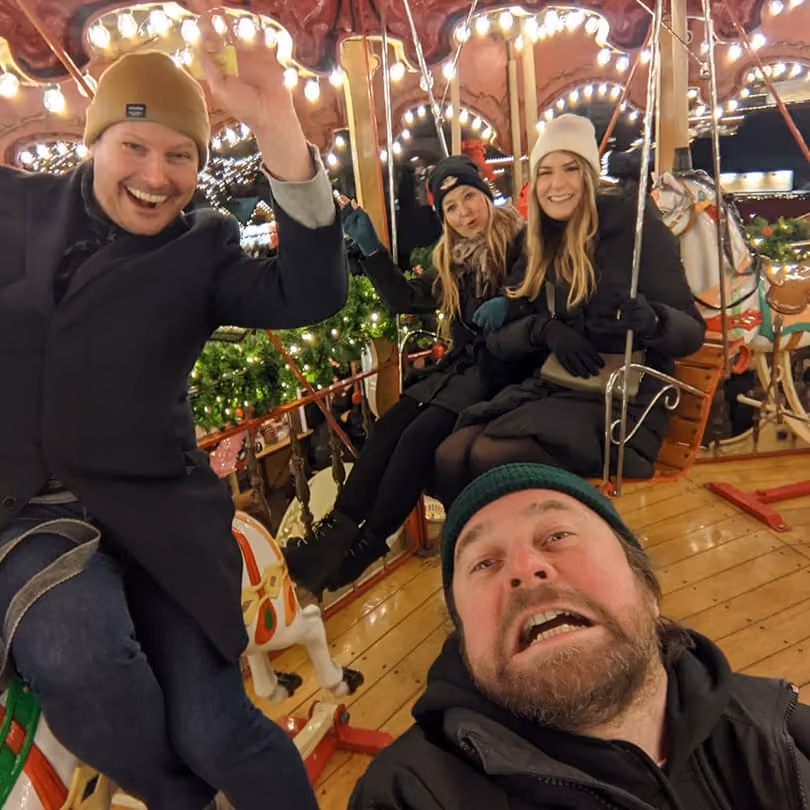 Four people smiling and waving on a brightly lit carousel with festive decorations.