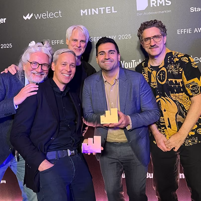 Five men smiling in front of a branded backdrop, two holding awards at an event.