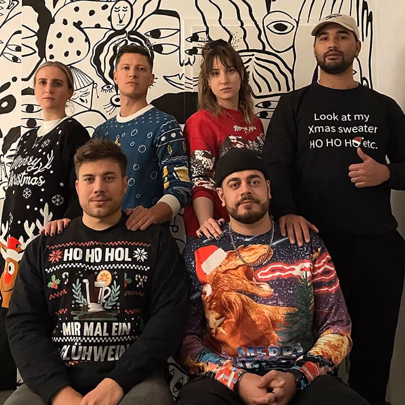 Six people wearing various holiday sweaters pose in front of a black and white abstract mural.