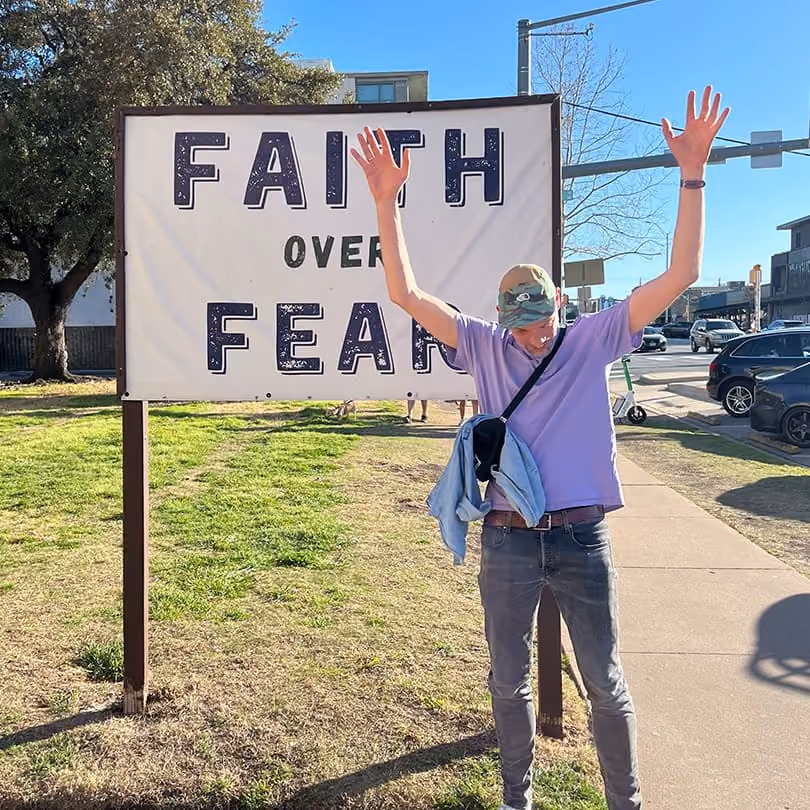 Person in casual clothes raising arms in front of a sign that says FAITH OVER FEAR.