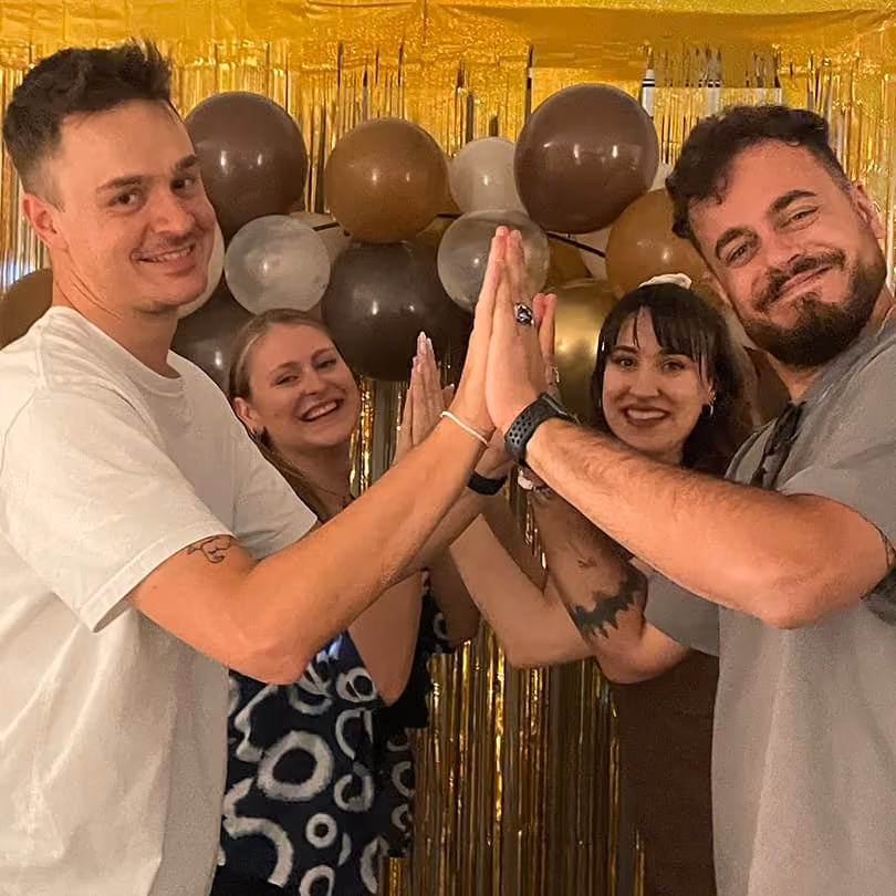 Four friends smiling and giving each other high fives at a party with brown and gold balloons behind.