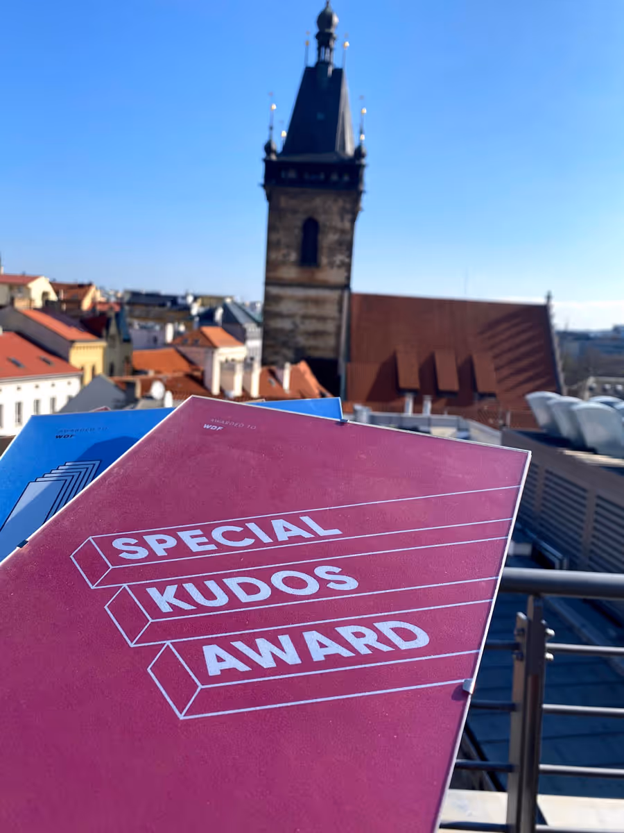 Magenta award booklet with the text 'Special Kudos Award' held up in front of a cityscape with a tall clock tower and rooftops under a clear blue sky.