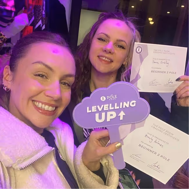 Two smiling women holding Beginner 2 Pole dancing certificates and a Levelling Up sign at The Pole Room.