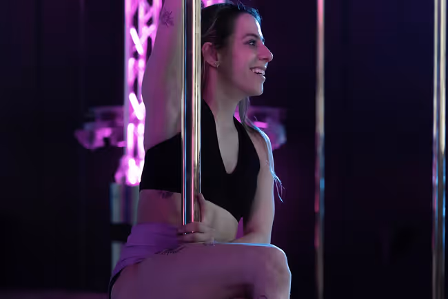 Woman smiling and posing on a dance pole under purple lighting in a dark studio.