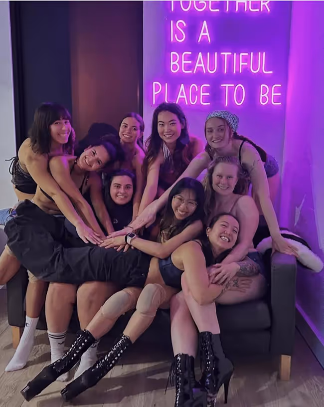 Group of nine young women smiling and sitting closely together on a couch under a neon sign that reads 'Together is a beautiful place to be.'